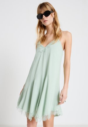 Frau trägt ein ärmelloses hellgrünes Kleid mit Spitzenbesatz und schwarze Sonnenbrille, steht vor einem einfarbigen weißen Hintergrund.