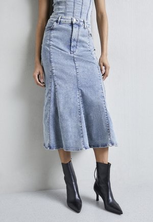 Kvinde iført en lyseblå denim midi nederdel og sorte læder spidse ankelstøvler med høje hæle, stående mod en lys væg.