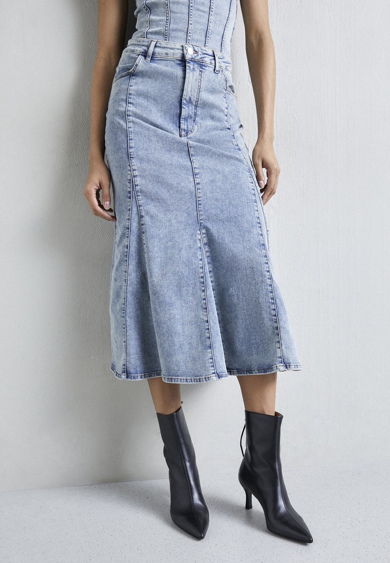 Femme portant une jupe midi en denim bleu clair et des bottines en cuir noir pointues à talons hauts, debout contre un mur clair.