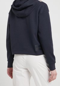 Mörkblå kort hoodie med textur och en dragsko i huvan. Har en logotyp-patch på baksidan och ribbade ärmslut för extra struktur.