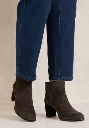 Personne portant des bottines en daim marron foncé à talons carrés et un jean bleu avec l'ourlet au-dessus des bottines.