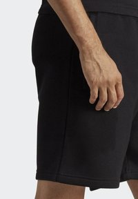 Pantaloni corti da allenamento neri con una texture liscia. Presentano tasche laterali e una vestibilità rilassata. Tessuto a presa manuale sui lati per dettaglio.