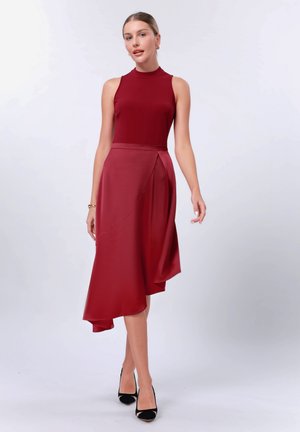 Red, sleeveless dress with a high neckline, asymmetrical hemline, and smooth fabric draping. Paired with black high-heel shoes.