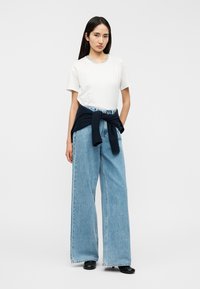 Maglietta bianca in cotone, jeans denim larghi azzurro chiaro, maglione blu navy annodato in vita, scarpe nere, con sfondo neutro.