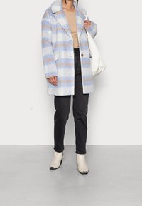 Blå og hvid ternet frakke med beige detaljer, knaplukning, stor krave, forlommer, båret over en rillet top og sorte jeans.