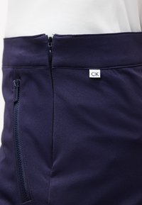 Κοντινό πλάνο στη μέση σκούρων μπλε παντελονιών με εμπρός φερμουάρ, πλαϊνή τσέπη με φερμουάρ και μικρή ετικέτα με το λογότυπο "CK" στη μέση.