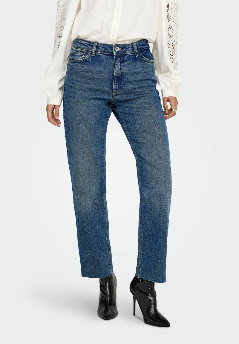 Jeans de mezclilla azul de cintura alta con pierna recta y dobladillo ligeramente deshilachado. Combinados con una blusa blanca de manga larga y botas de tacón negras.