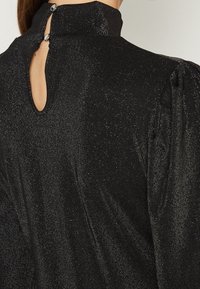 Svart klänning med hög krage, textil med struktur som inkluderar metalltrådar, och en nyckelhålsdetalj på baksidan som säkras med två knappar.