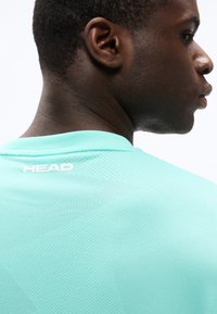 Νεαρός άνδρας που φοράει ανοιχτό μπλε σπορ πουκάμισο με τρύπες, με το λογότυπο "HEAD" εμφανές στο πάνω μέρος της πλάτης, κοιτάζοντας προς τα πλάγια.