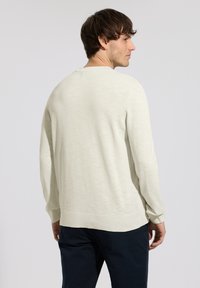 Homme de dos portant un pull à manches longues de couleur crème et un pantalon foncé, sur un fond clair uni.