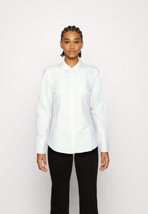 Camisa blanca de botones con mangas largas, cuello clásico, textura suave y un ligero brillo; combinada con pantalones negros acampanados.