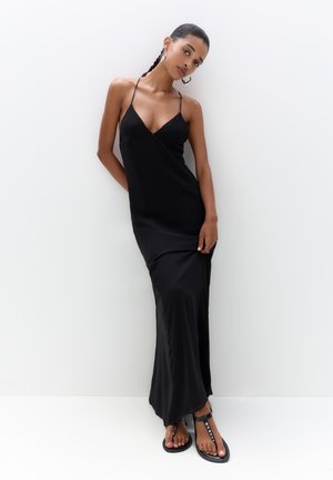 Schwarzes Maxikleid mit V-Ausschnitt, dünnen Trägern und einem fließenden Design, ergänzt durch flache schwarze Sandalen mit dekorativen Hardware-Akzenten.