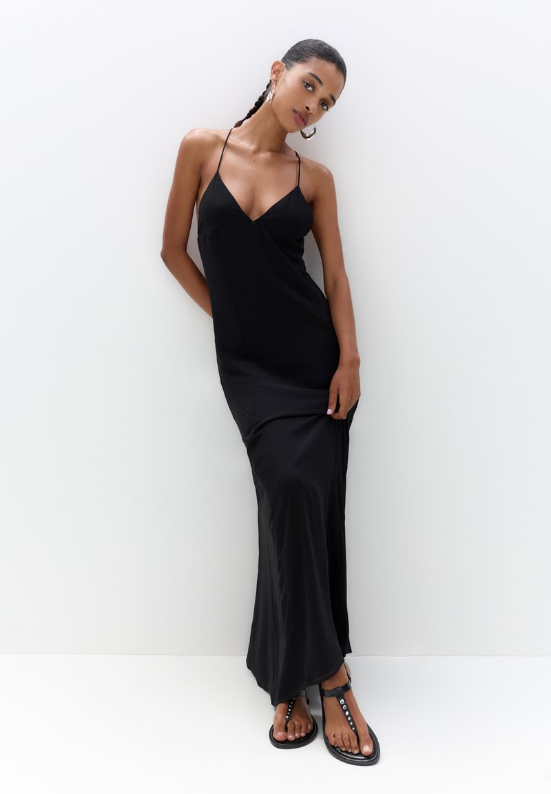 Schwarzes Maxikleid mit V-Ausschnitt, dünnen Trägern und einem fließenden Design, ergänzt durch flache schwarze Sandalen mit dekorativen Hardware-Akzenten.