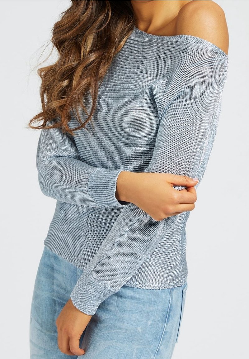 Femme portant un pull tricoté bleu clair sans épaules et un jean en denim, avec de longs cheveux bruns ondulés, bras croisés sur le corps.