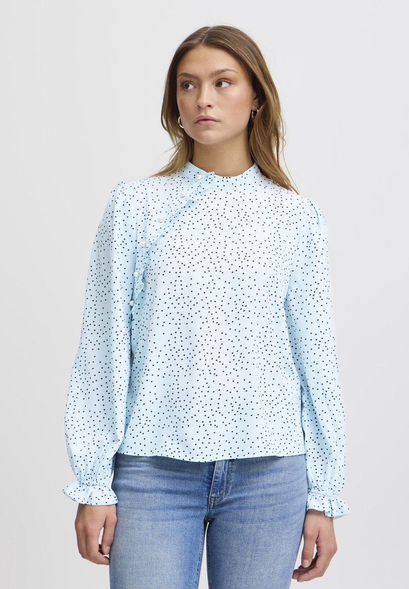 Blusa azzurro chiaro con pois neri, dettaglio arricciato lungo la spalla sinistra e maniche a sbuffo con polsini elasticizzati.