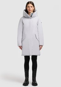 Šviesiai pilka, vidutinio ilgio pūstas paltas su plačia kapišonu, priekiu su užtrauktuku ir dviem šoninėmis kišenėmis, derinamas su juodomis tamprėmis ir stambiais juodais batais.