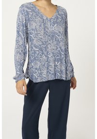 Blusa blu a maniche lunghe con motivo foglia bianca, scollatura a V e polsini arricciati. Abbinata a pantaloni blu scuro. Tessuto morbido e leggero.