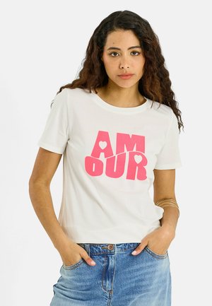 Mujer con cabello largo y rizado lleva una camiseta blanca con texto rosa "AMOUR" y vaqueros azules, manos en los bolsillos, expresión neutral, fondo blanco.
