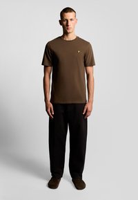 T-shirt marrone a maniche corte con collo rotondo, caratterizzato da un piccolo logo giallo sul petto. Abbinato a pantaloni neri larghi e ciabatte scure.