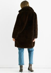 Brown faux fur coat with a high collar and long sleeves, featuring a relaxed fit, and worn over light blue jeans and patterned ankle boots.