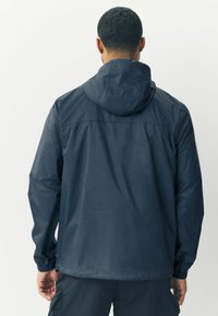 Muž v tmavě modré větrovce s kapucí a cargo kalhotách, stojící zády k jednoduchému světlému pozadí.