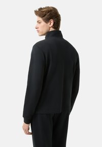 Czarny pulower z wysokim kołnierzem, wykonany z gładkiej tkaniny. Posiada luźny krój i prążkowane mankiety, widziany od tyłu.