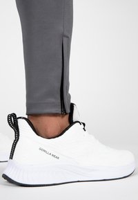 Atletiek schoenen in het wit met een gestructureerde mesh bovenkant, zwarte accenten en een merknaam aan de zijkant; gecombineerd met grijze trainingsbroek met zijritsen.