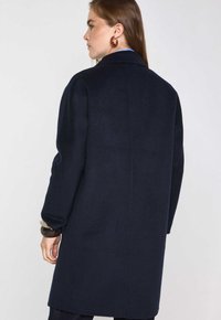 Cappotto di lana blu navy con un design strutturato, maniche lunghe e taglio dritto. Presenta un colletto classico e una texture liscia.