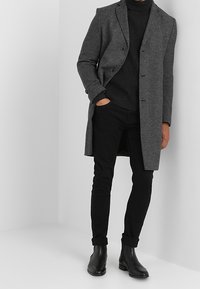 Manteau gris à motifs avec un design croisé, porté sur un col roulé noir et un pantalon slim noir, associé à des bottines noires.
