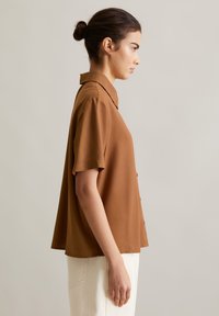 Bruine korte mouwen shirt met een kraag, knoopsluiting en een zachte textuur. Gepaard met lichtgekleurde broek, van opzij weergegeven.