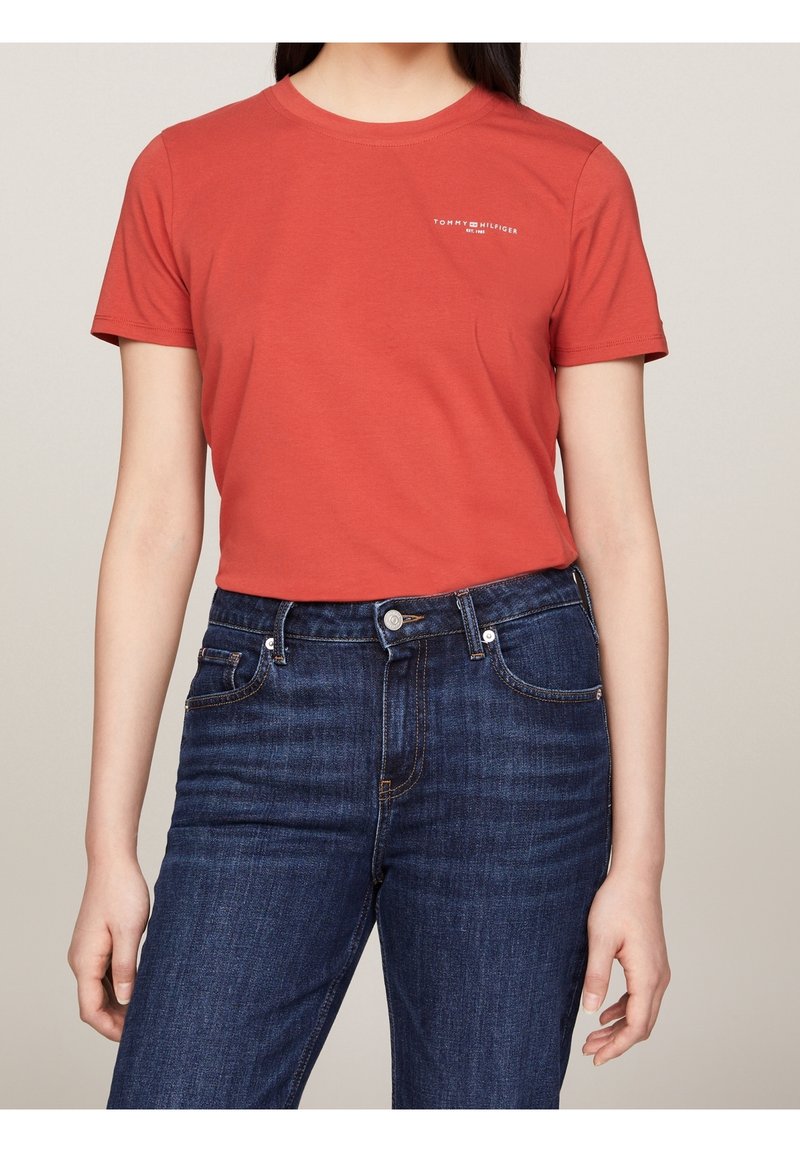 Rød bomulds T-shirt med rund hals og korte ærmer; har et hvidt logo på brystet. Båret med mørkeblå denim jeans.
