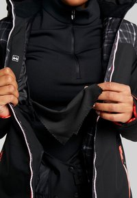 Schwarze Jacke mit hohem Kragen, kontrastierenden roten Akzenten und kariertem Innenfutter. Zeigt ein weiches schwarzes Stoffmuster innen.