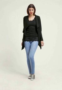 Vrouw staat in een zwart vest, zwart top met kant, lichtblauwe skinny jeans en grijze sneakers tegen een effen achtergrond.