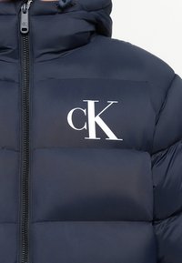 Navyblauwe pufferjack met een ritssluiting aan de voorkant en een wit "cK" logo op de linkerborst.