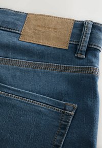 Vaqueros de mezclilla azul oscuro con una textura suave, que presentan una etiqueta de cuero marrón marcada con "DENIM SUPPLY" y detalles de costura en contraste.