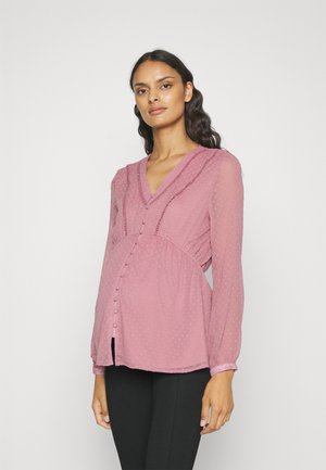 Maternity blouse in light pink, sheer fabric with textured dots, ruffled neckline, button detail, and long sleeves. Fitted under the bust.