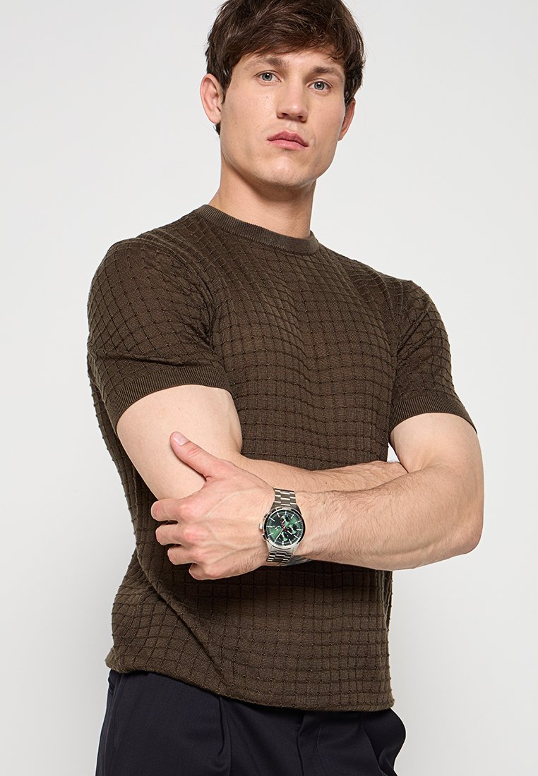 Jersey corto de manga corta de punto texturizado en color marrón con cuello redondo. Se lleva con un reloj de tono plateado con esfera verde y correa metálica.