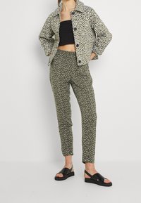 Zielona kurtka pikowana z białym wzorem kwiatowym, czarny top na krótki rękaw oraz oliwkowe spodnie w wzory, zestawione z czarnymi sandałami, stojąc na neutralnym tle.