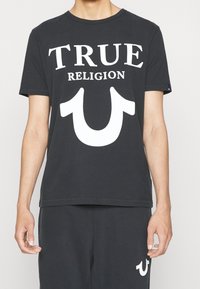T-shirt noir en coton avec col rond, arborant le texte blanc "TRUE RELIGION" et un grand logo en forme de fer à cheval blanc sur le devant.