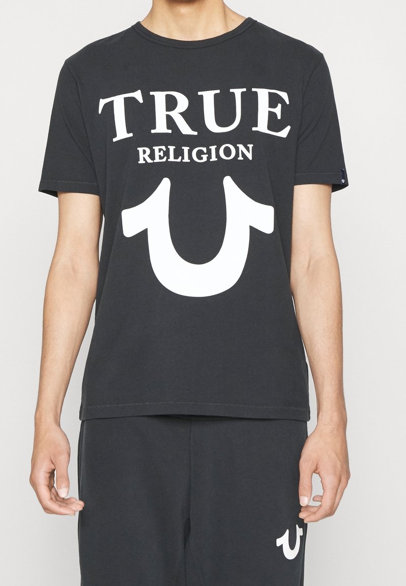 T-shirt noir en coton avec col rond, arborant le texte blanc "TRUE RELIGION" et un grand logo en forme de fer à cheval blanc sur le devant.