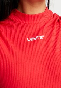 Červený žebrovaný top s vysokým límcem a vytlačeným bílým logem "LeVi's" na hrudi. Hladká textura, přiléhavý střih, casual styl.