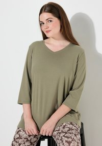 Olijfgroene V-hals t-shirt met 3/4 mouwen, gemaakt van zachte stof. Draag over bruine leggings met een patroon van bloem ontwerpen.