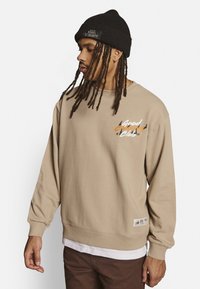 Beige sweatshirts med rund hals og lange ærmer, prydet med et farverigt grafisk tryk "Good Company". Båret over en hvid skjorte, med en tekstureret strikket hue.