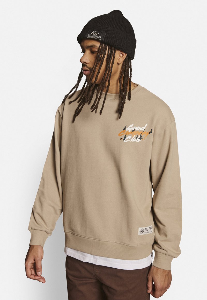 Beige Crewneck-Sweatshirt mit langen Ärmeln, ausgestattet mit einem bunten grafischen "Good Company"-Druck. Trägt man über einem weißen Hemd und einer strukturierten Strickmütze.