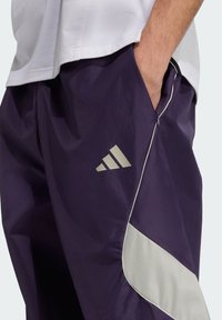 Fialové športové nohavice s lesklým povrchom, s šedým svetlým lemovaním a logom Adidas. Navrhnuté so bočnými vreckami pre väčšiu funkčnosť.
