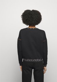 Persona con pelo rizado que lleva una camiseta de manga larga negra con puños y dobladillo con patrones, de pie frente a una pared blanca lisa.