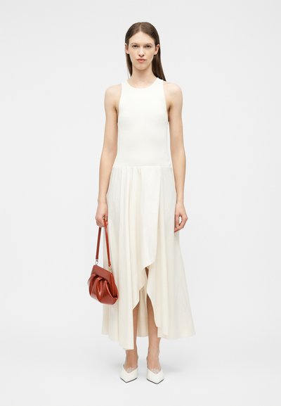 Γυναίκα που φοράει αμάνικο λευκό φόρεμα με ασύμμετρη τελειώση, κρατώντας μικρή καφέ τσάντα και φορώντας λευκά μυτερά παπούτσια.