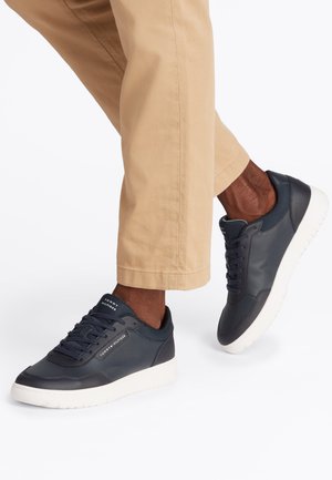 Mann trägt beige Hose und marineblaue Tommy Hilfiger Sneaker mit weißen Sohlen, macht einen Schritt nach vorne vor weißem Hintergrund.