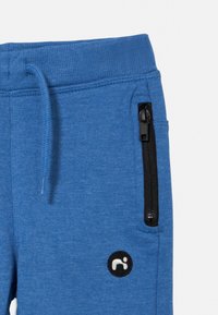 Kék melegítőnadrág puha anyagból, oldalt fekete cipzáras zsebbel és egy kis fekete-fehér márkajelzéssel.