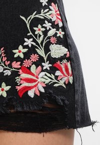 Shorts en denim noir avec ourlet effiloché, ornés d'une broderie florale en tons rouges, roses et verts sur un fond texturé.